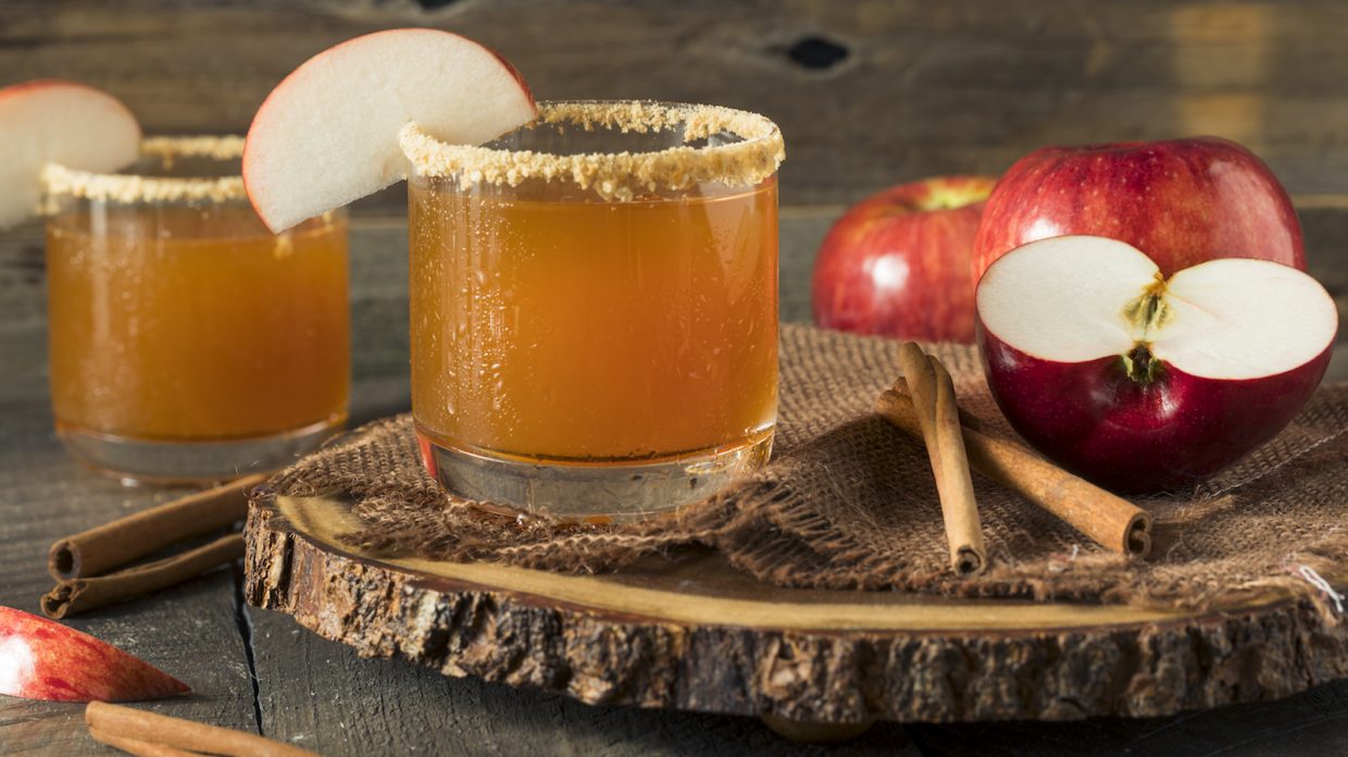 brown cocktail in rocks glass with a slice of apple on the rim and cinnamon sticks and apples in the background