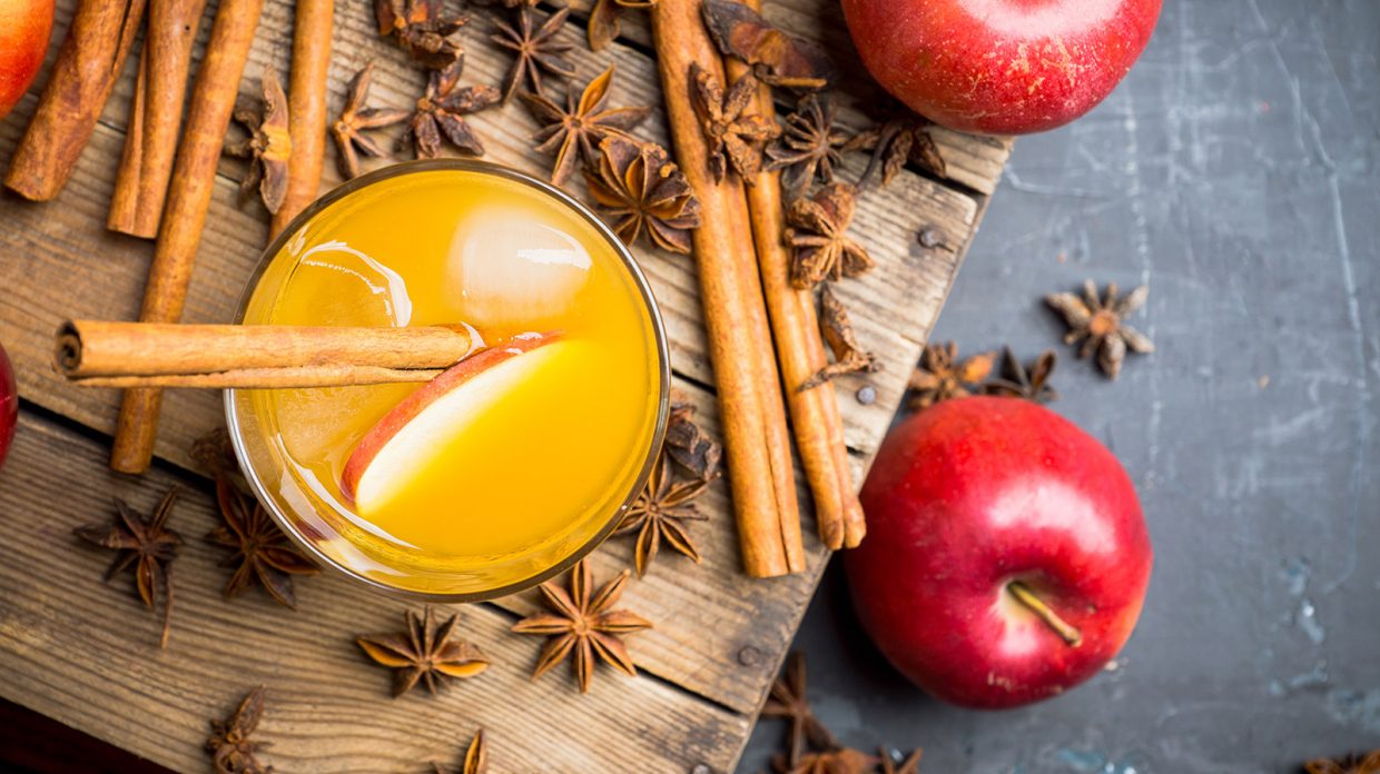 A cocktail garnished with a cinnamon stick and apple slice resembling the Brandied Rye and Apple Punch cocktail rests on a wooden board next to more apples and cinnamon sticks.