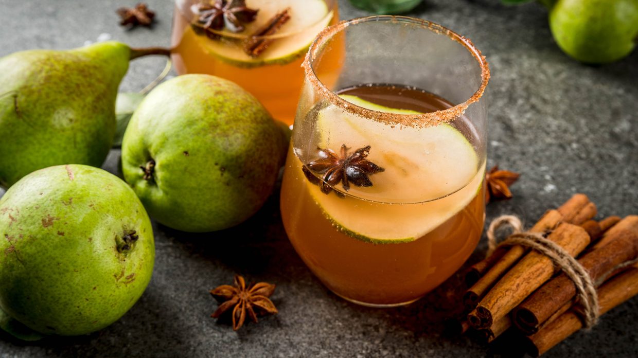 A cocktail garnished with a pear slice and spices, alongside a bundle of cinnamon sticks, pears, and star anise, resembling the Bucky Barnes cocktail rests upon a gray surface.