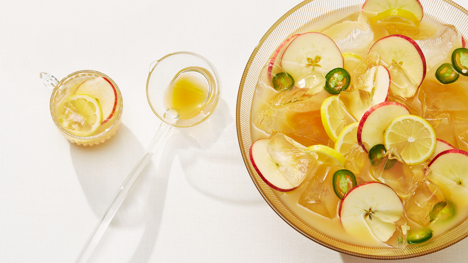 A bowl of Galway Cider whisky punch, garnished with apple, jalapeño, and lemon slices, rests on a white surface next to two punch-filled glass cups with a serving spoon.