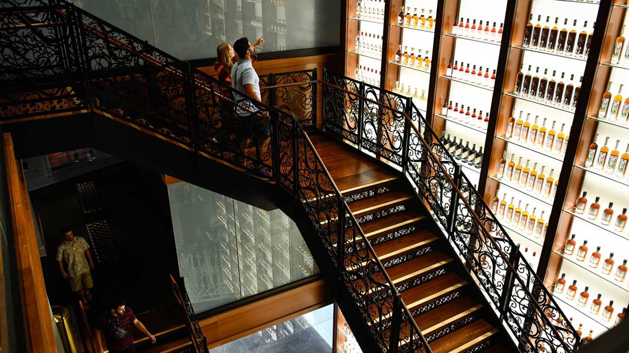 Visitors at the newly opened Sazerac House cocktail museum in New Orleans descend a staircase past a three-story bottle wall showcasing various Sazerac spirits.