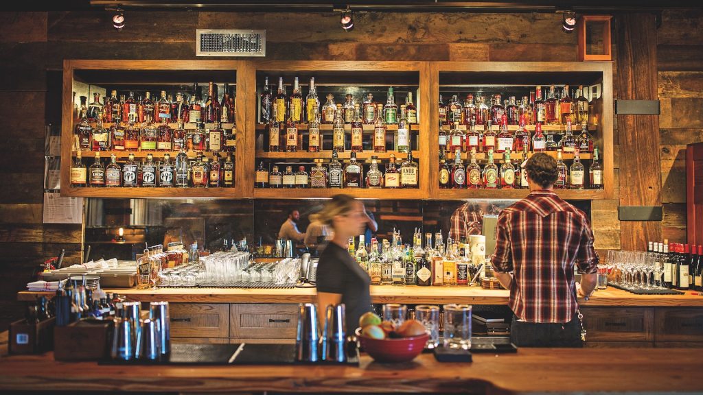 A man and woman stand behind a bar, facing a vast amount of alcohol bottles.