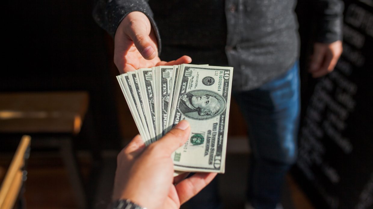 A man hands another man a large stack of ten-dollar bills.