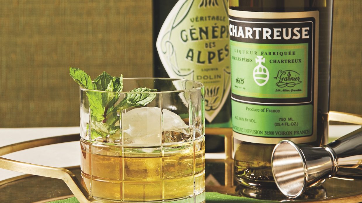 A cocktail in a rocks glass and garnished with a mint sprig sits in front of two bottles of chartreuse.