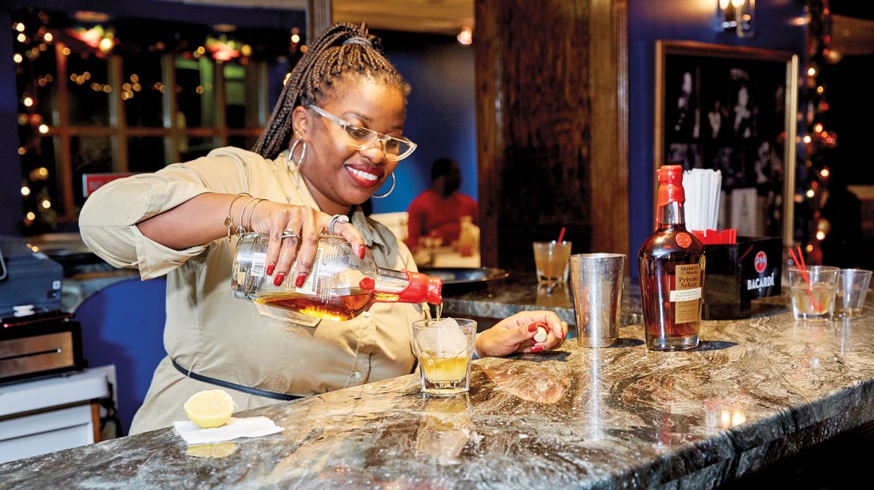 A woman wearing red lipstick and nail polish, hoop earrings and bracelets, her braided hair in a ponytail, pours from what appears to be a bottle of Maker's Mark Private Select bourbon over a large ice cube in a rocks glass resting atop what appears to be a marble bar at Sylvia's Restaurant in Harlem on Dec. 5, 2019.