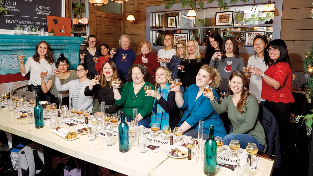 Women Who Whiskey founder and principal Julia Ritz Toffoli, standing, clad in a white scalloped-edge sweater, raises a glass, along with other club members, some standing, others seated at a long table laden with whisky flights and various snacks, at Tara Rose bar in New York City on Dec. 16, 2019.