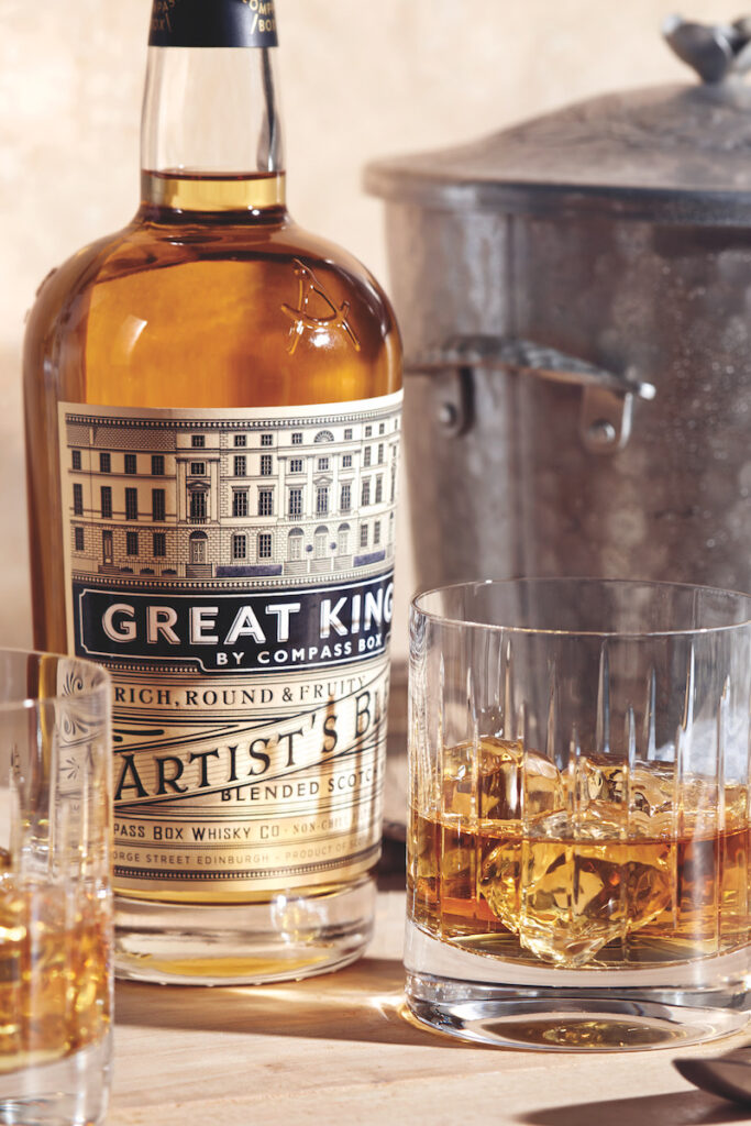 A bottle of Compass Box Great King Street Artist's Blend rests atop a wooden surface along with a bar spoon and two rocks glasses containing ice and what appears to be whisky, and in the background, a metal ice bucket ice bucket with a lid and small handles.