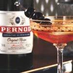 The auburn-hued Cocktail à la Louisiane, in a fancy chilled coupe, garnished with three speared maraschino cherries, rests on a dark tray, a bottle of Pernod Original Recipe absinthe visible in the background.