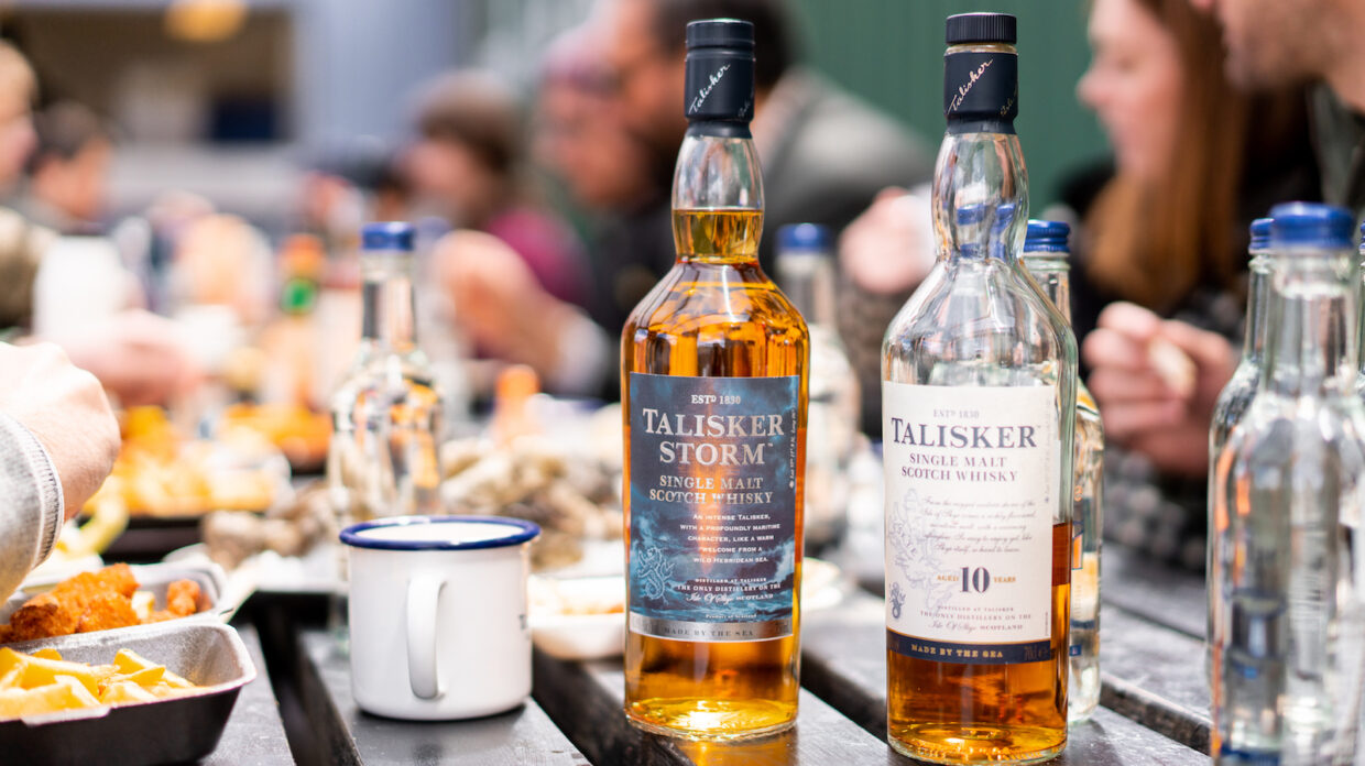 Two bottles of Talisker scotch rest atop a table around which people sit eating oysters and drinking whisky.