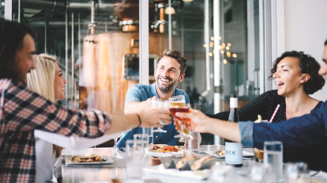 A group of young people clink glasses at Bottle & Bond Kitchen & Bar, with distillery equipment in the background