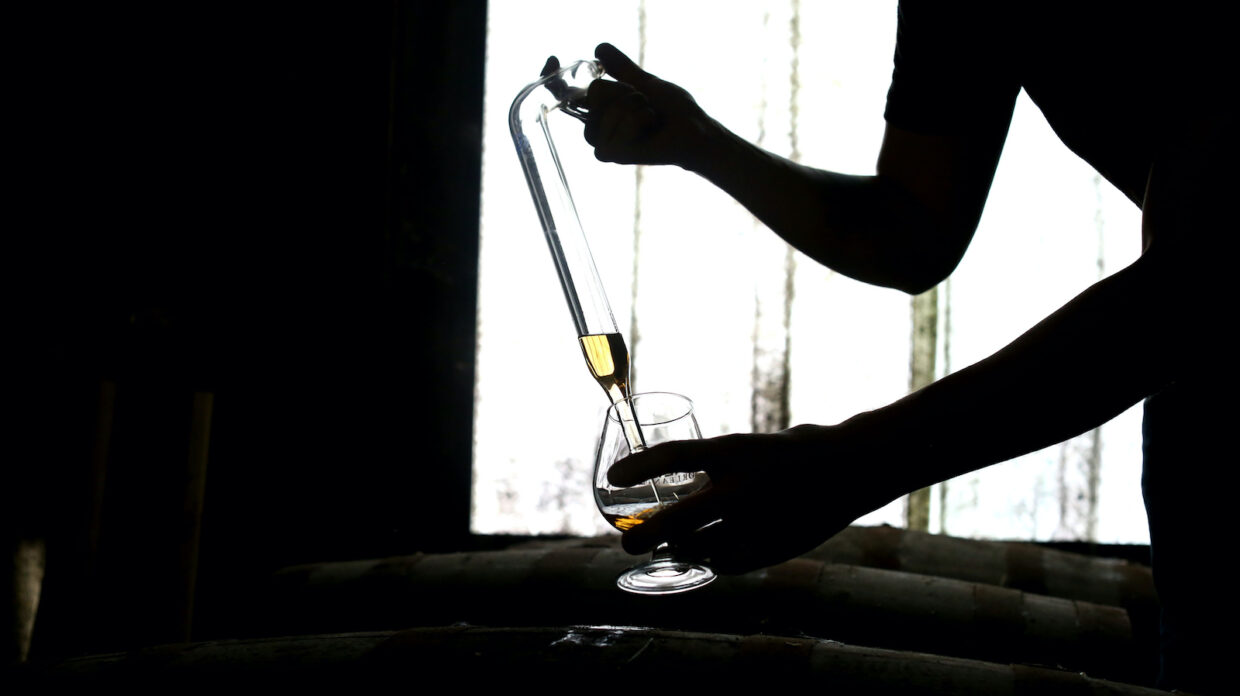 silhouette of a person using a barrel thief to sample amber liquid from a cask and releasing it into a balloon glass