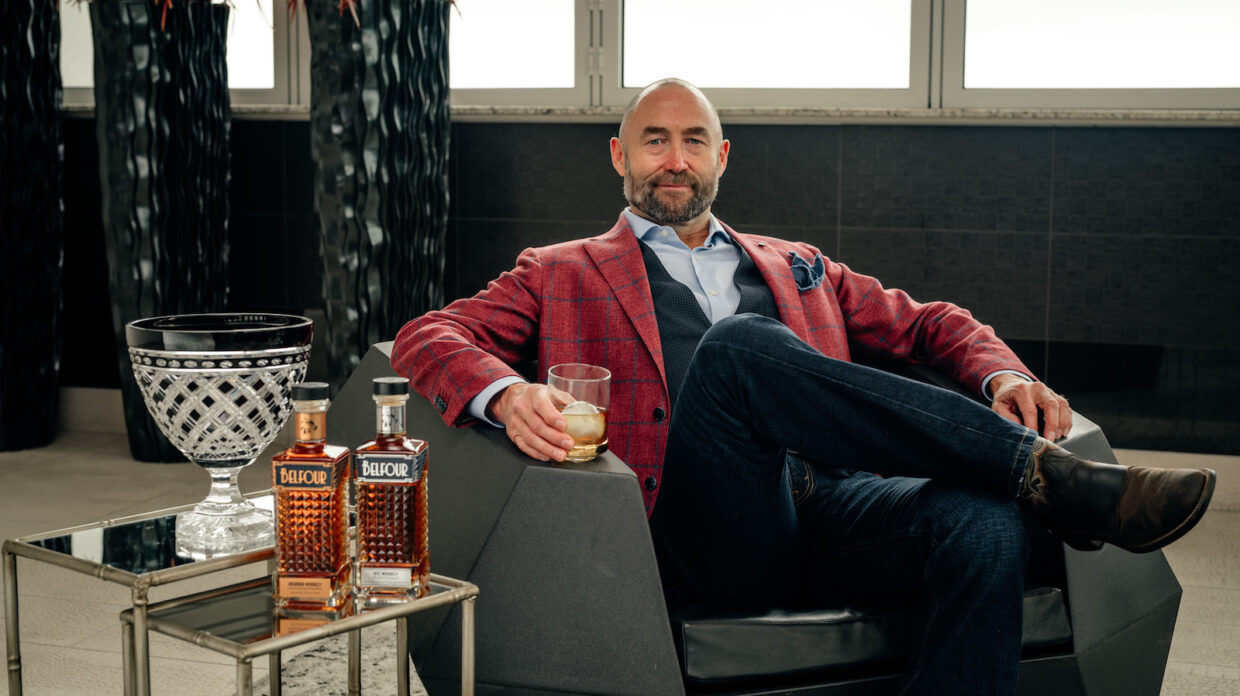 A man sits in a lounger chair holding a glass of whiskey. Two whiskey bottles sit on the table near him.