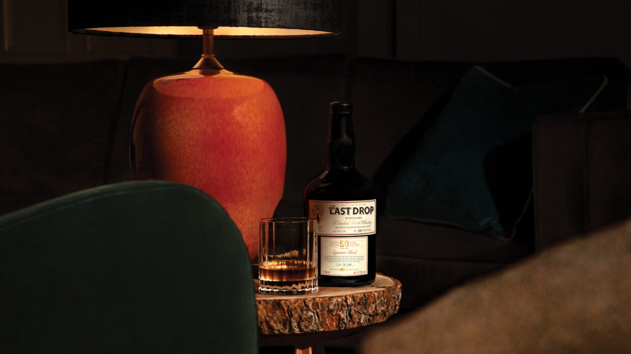 A bottle of whisky and glass of whisky sit on a table under low light.