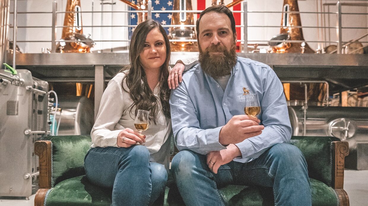 A man and a woman pose with a glass of whiskey in front of stills